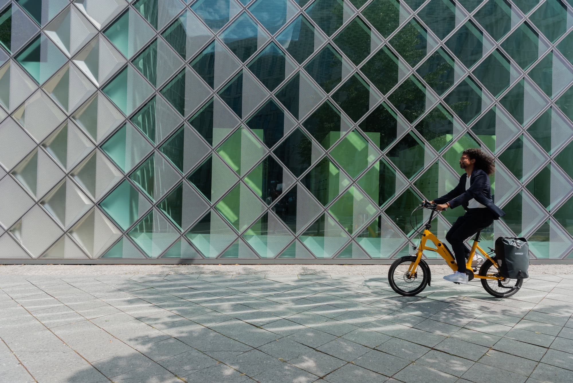 Person fährt Fahrrad vor moderner Glasfassade; lächelnd, gelbes Fahrrad, geometrische Muster.