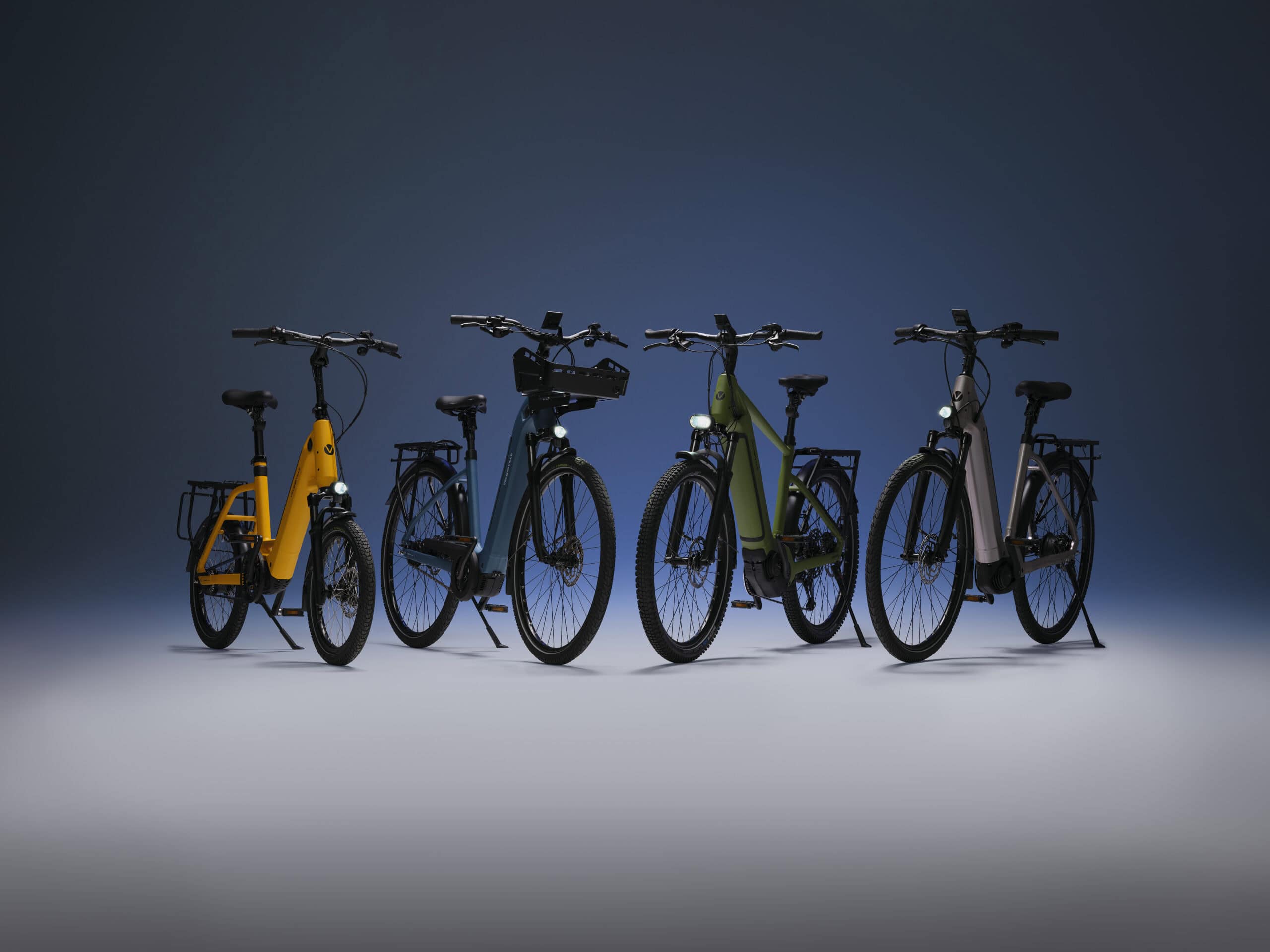 Vier moderne E-Bikes in unterschiedlichen Farben stehen aufgereiht vor einem dunklen Hintergrund.